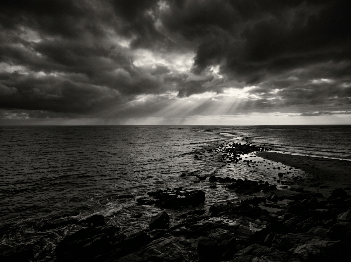 Rocky shoreline and ocean beneath storm clouds with rays of sunlight
