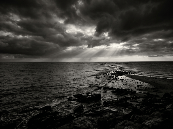 Rocky shoreline and ocean beneath storm clouds with rays of sunlight