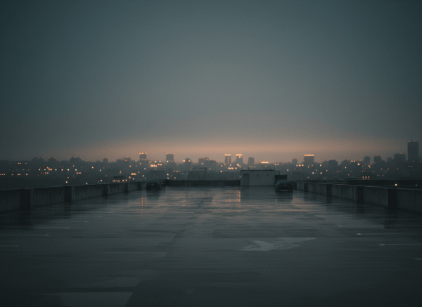 A minimalistic, subtly moody scene: a rain-wet rooftop parking lot at dusk, near-empty, with faint city lights glowing in the distance. Desaturated tones — deep grays, muted amber from distant streetlights. No people, no text. Cinematic, quiet, introspective. 4:3 aspect ratio.